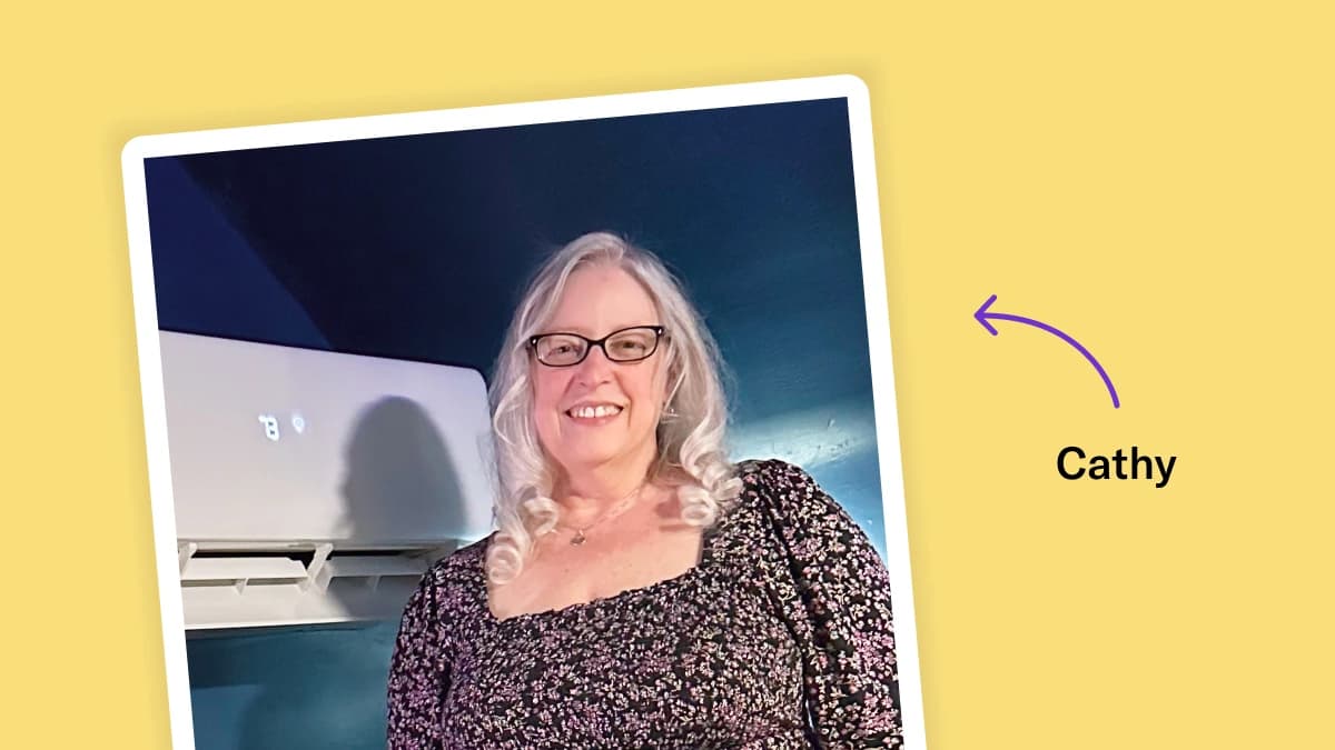 Polaroid-style photo of Cathy on a yellow background. Cathy is smiling, wears glasses, has curled white hair, and wears a square neck printed floral top. She stands next to her mini-split inside of a room painted blue.