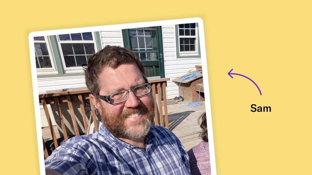 Selfie of Sam Ose in front of his house on the deck. He is smiling and has short brown hair, a short beard, and wears glasses.