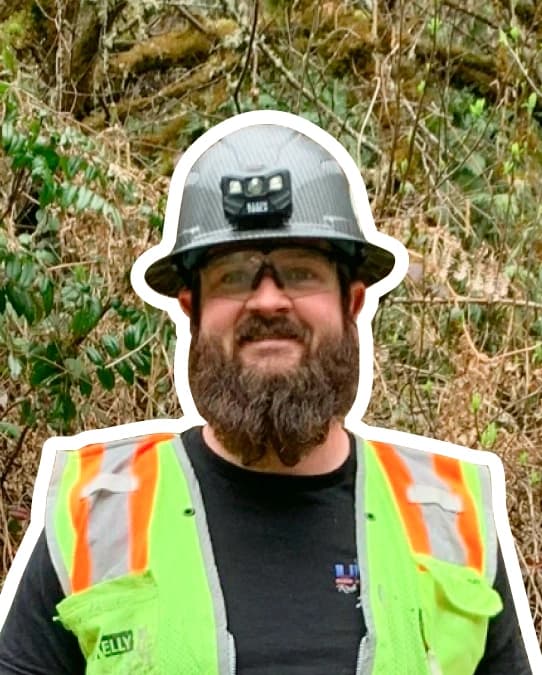White outlined photo of Ryan Smith standing in front of forest trees and brush. Ryan Smith wears a gray hard hat, safety glasses, a grey long sleeve shirt, and a bright green high vis vest.