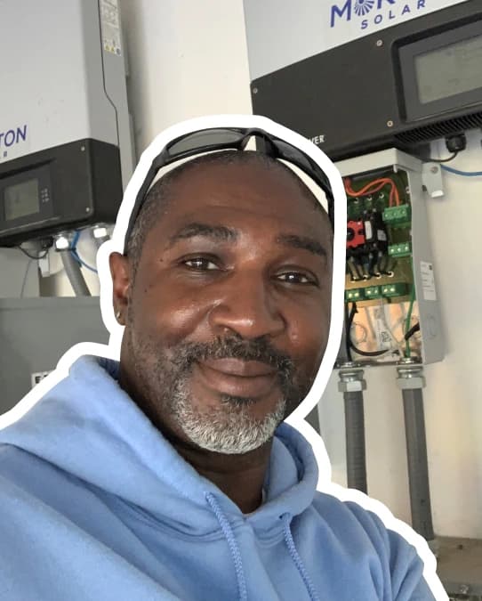White outlined photo of Monquale Owsley in front of a solar inverter. He is smiling at the camera, has gray facial hair, is wearing a light blue hoodie and safety glasses on the top of his head.