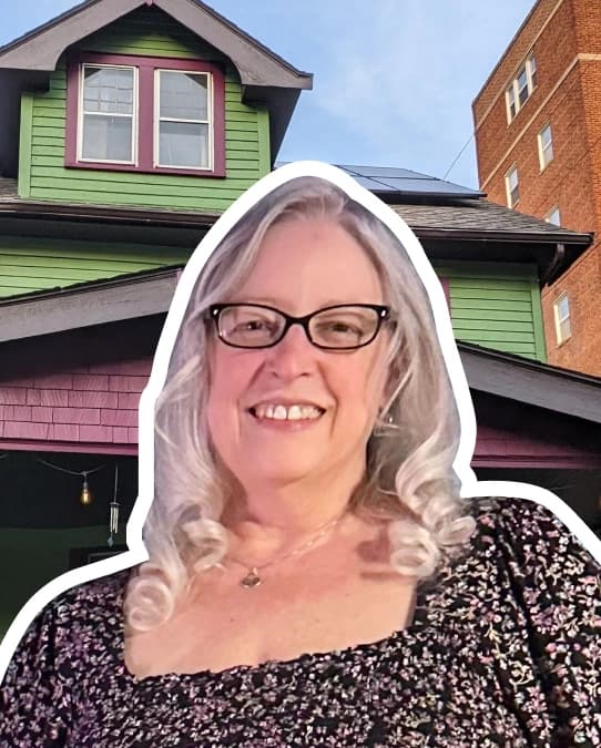 White outlined photo of Cathy, who is smiling, wears glasses, has curled white hair, and wears a square neck printed floral top. Her photo is on top of an image of her house, which is painted green and purple-magenta on the exterior.